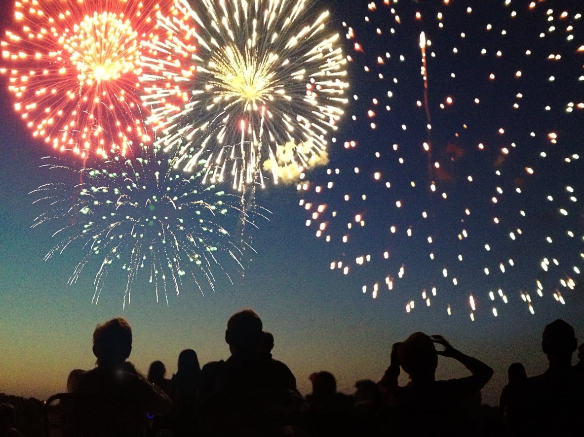A Fourth of July fireworks display exploding in the sky over a group of people. The crowd, spectators in silhouette, celebrate the Independence Day national holiday together, looking up and watching bursts of color. The pyrotechnic performance may represent New Year’s Eve, Chinese New Year, or other traditional festival celebration events.