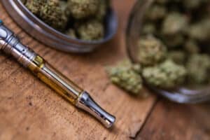 THC oil concentrate filled vape pen on natural wood with an open glass container full of Mango Kush strain marijuana buds grown & sold in dispensaries through-out Southern California.