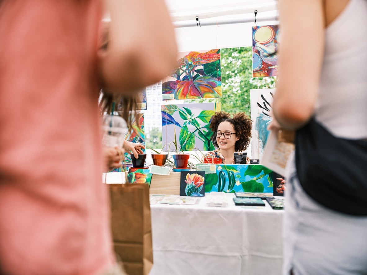 Young Latin woman artist selling her art at Outdoor Market. She is dressed in casual East Indian dress. Exterior of public park in downtown Toronto, Canada.